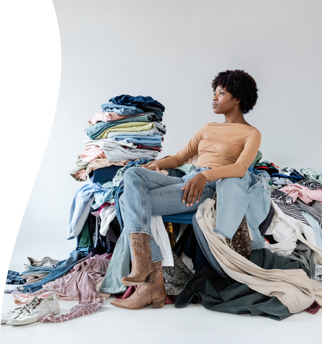 An African woman with short afro hair sits comfortably atop a massive, overflowing pile of colorful clothing against a plain white studio background. She wears a beige long-sleeved top, light blue jeans, and light brown cowboy boots, gazing pensively off to the side. Neat stacks of folded garments rest near her shoulder, while loose shirts, pants, sweaters, and shoes spill across the floor around her.